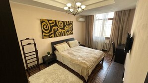 A bedroom with a large bed, a nightstand, a chair, a desk, and a wall-mounted TV.