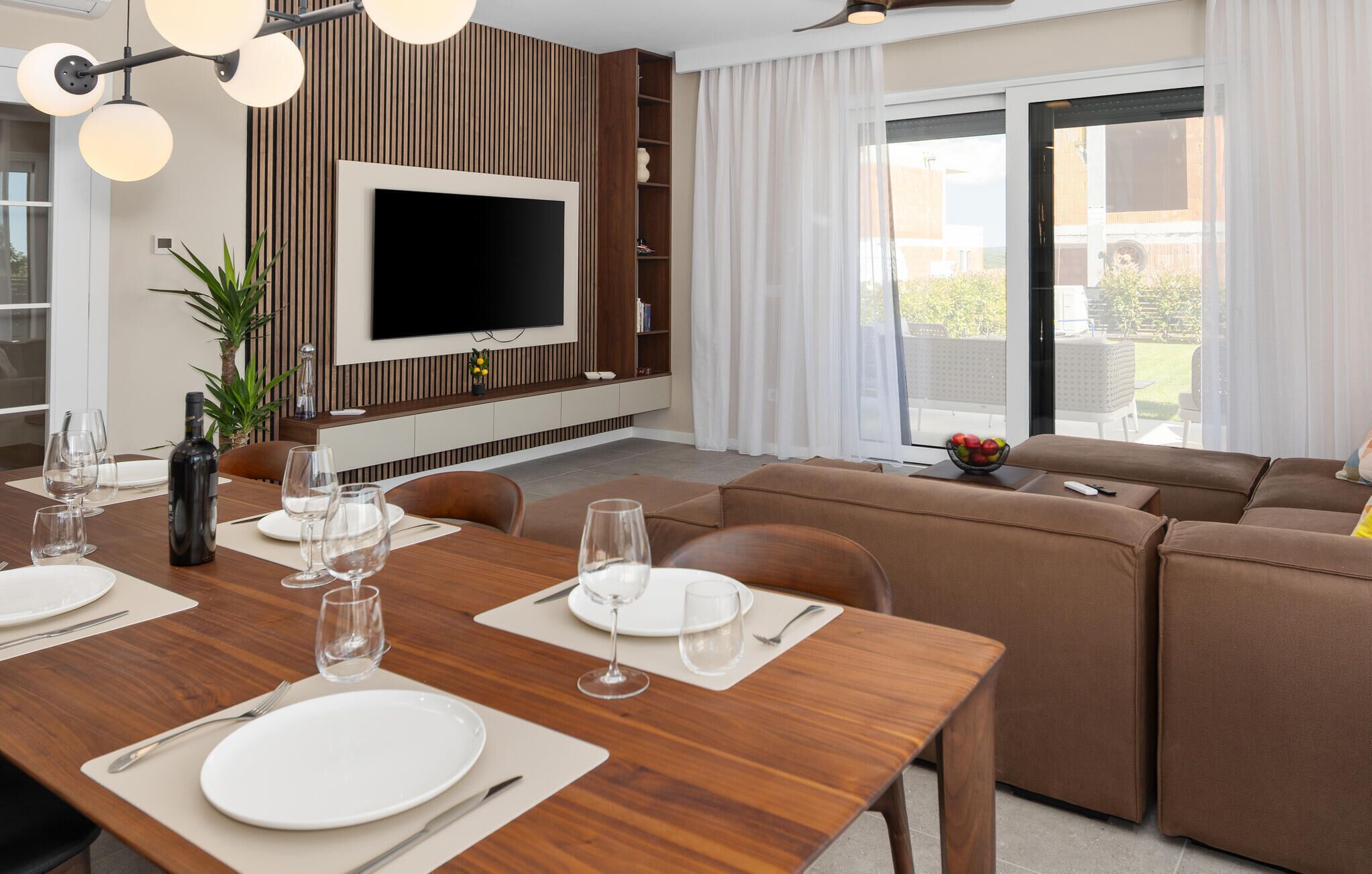A modern living room with a dining area, a flat-screen TV mounted on a wooden wall, and a large sliding glass door leading to an outdoor area.