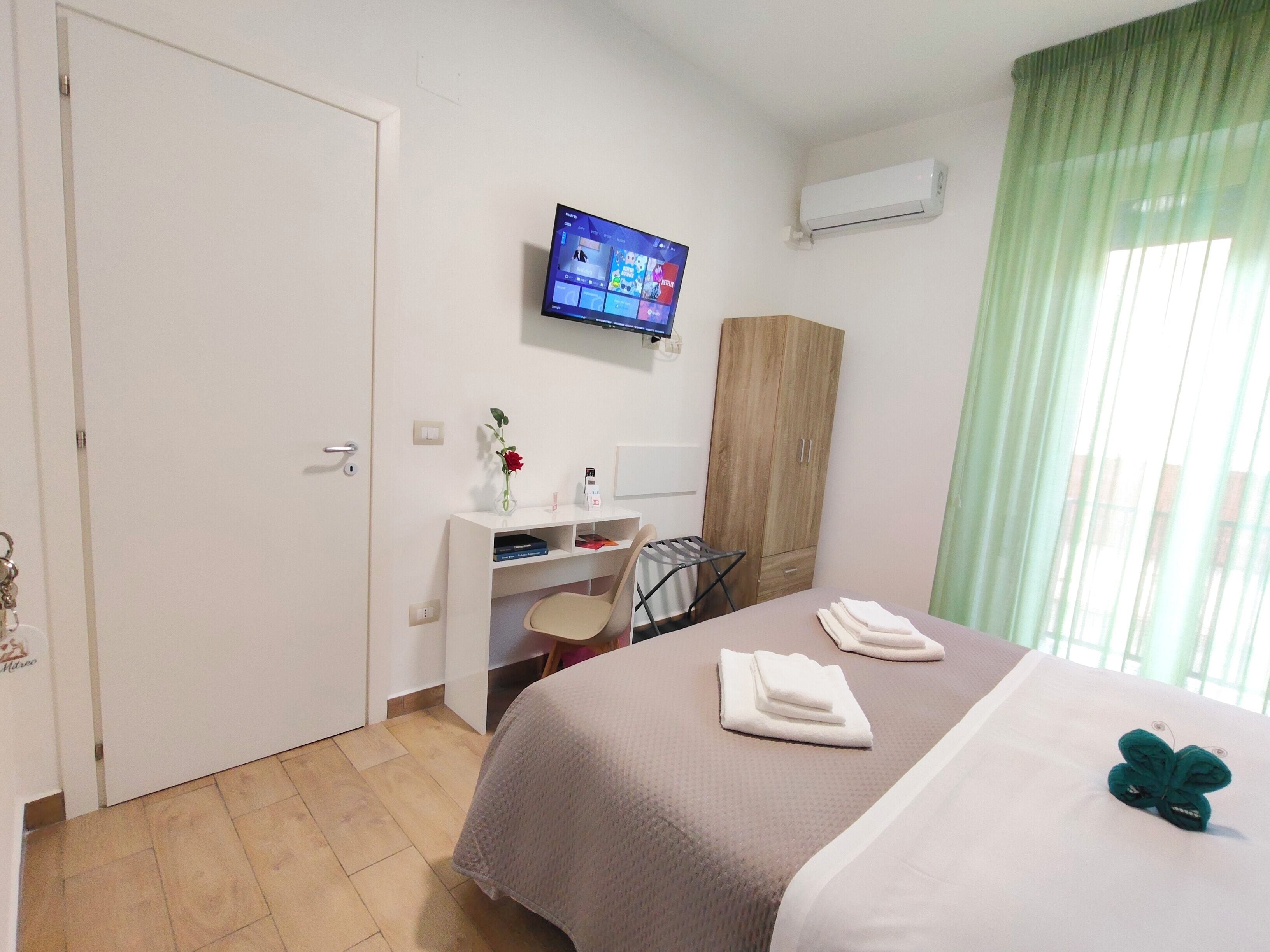 A hotel room with a bed, a desk with a chair, a television mounted on the wall, and a wardrobe.