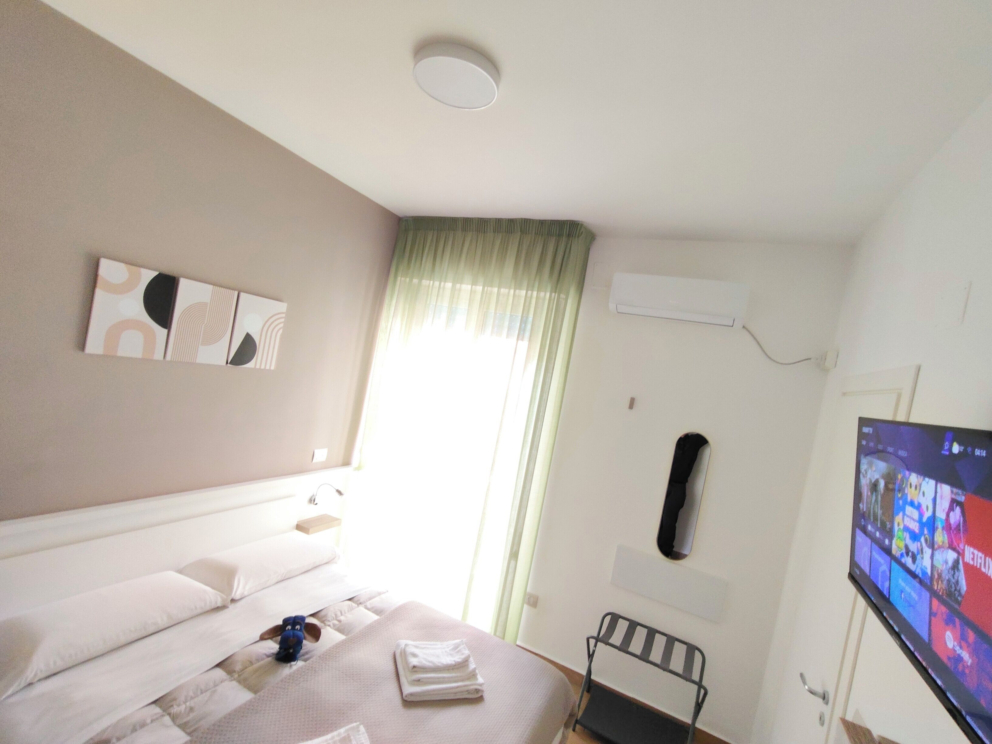 A hotel room with a bed, a TV, a window with curtains, and a wall-mounted air conditioner.