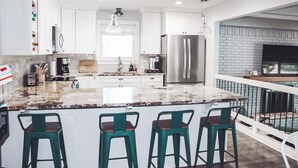 Una cucina moderna con mobili bianchi, una grande isola con sgabelli da bar, elettrodomestici in acciaio inossidabile e un lampadario.