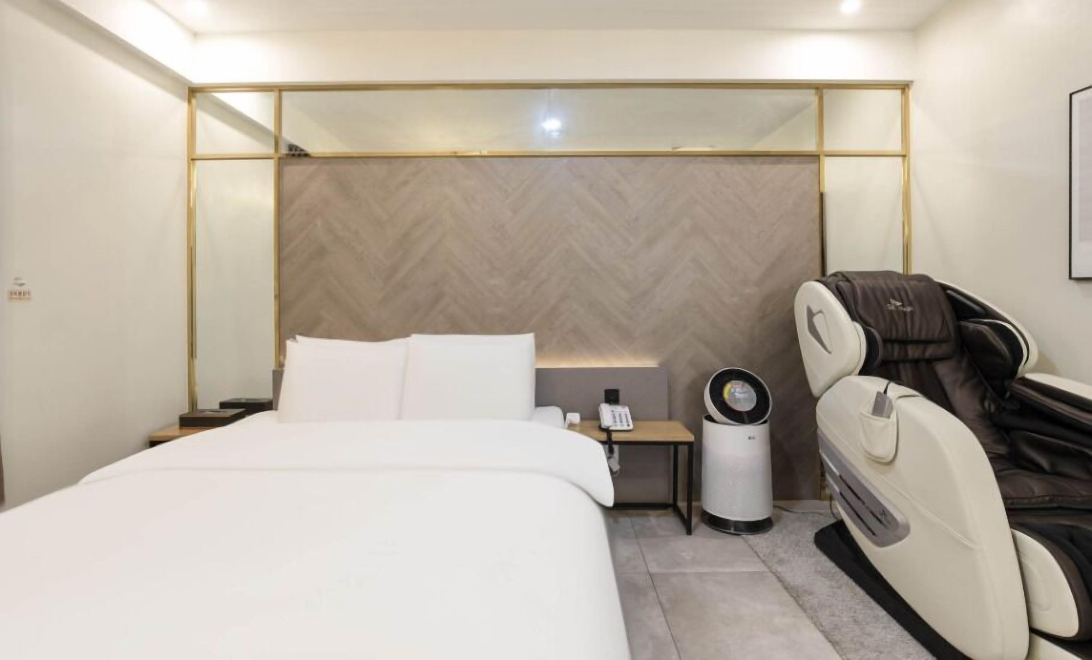 A hotel room with a bed, a massage chair, a bedside table with a phone, and a trash bin.