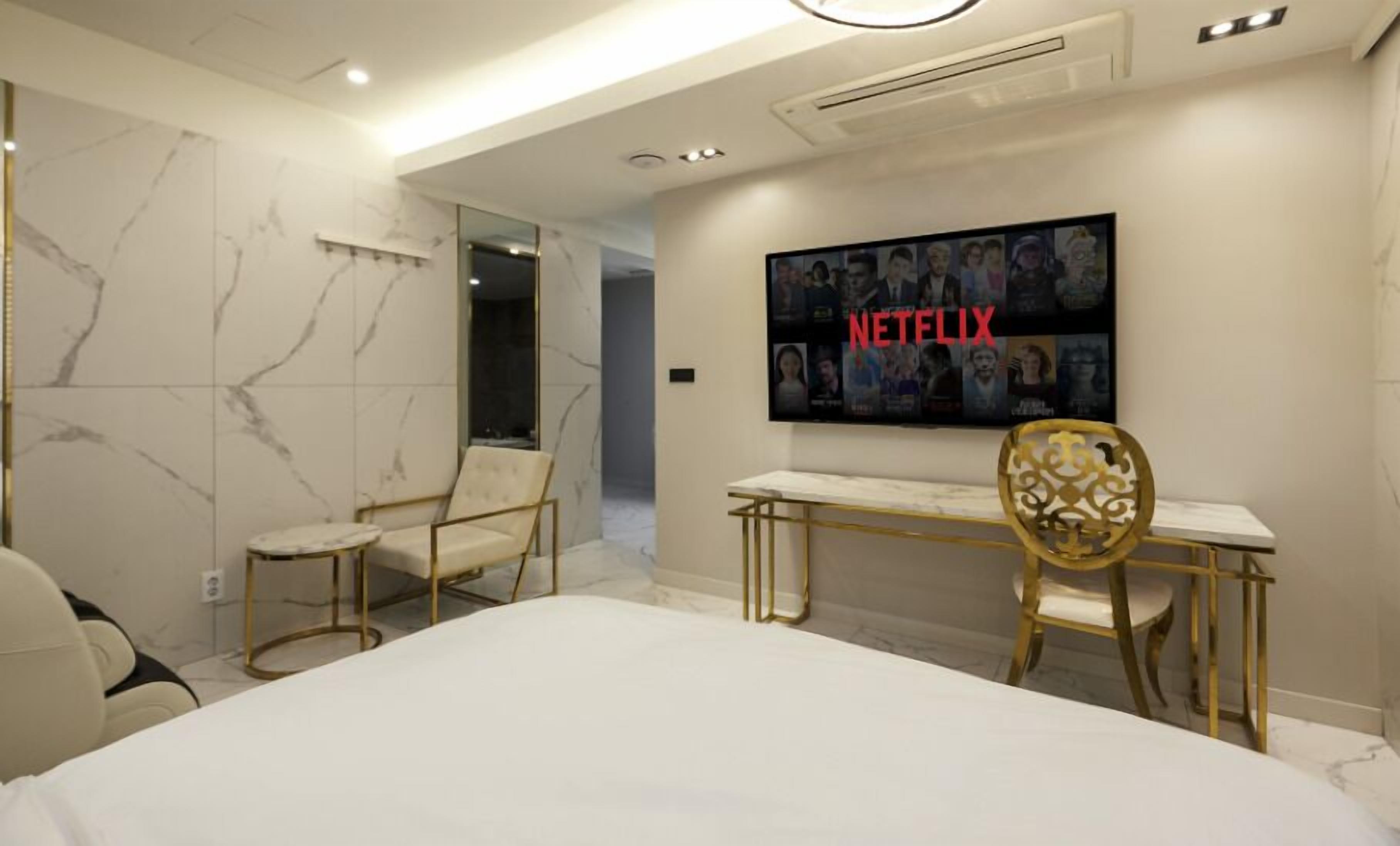 A modern hotel room with a large bed, a desk, and a chair. There is a television mounted on the wall displaying the Netflix logo.