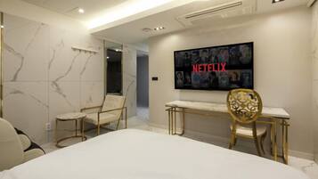 A modern hotel room with a large bed, a desk, and a chair. There is a television mounted on the wall displaying the Netflix logo.