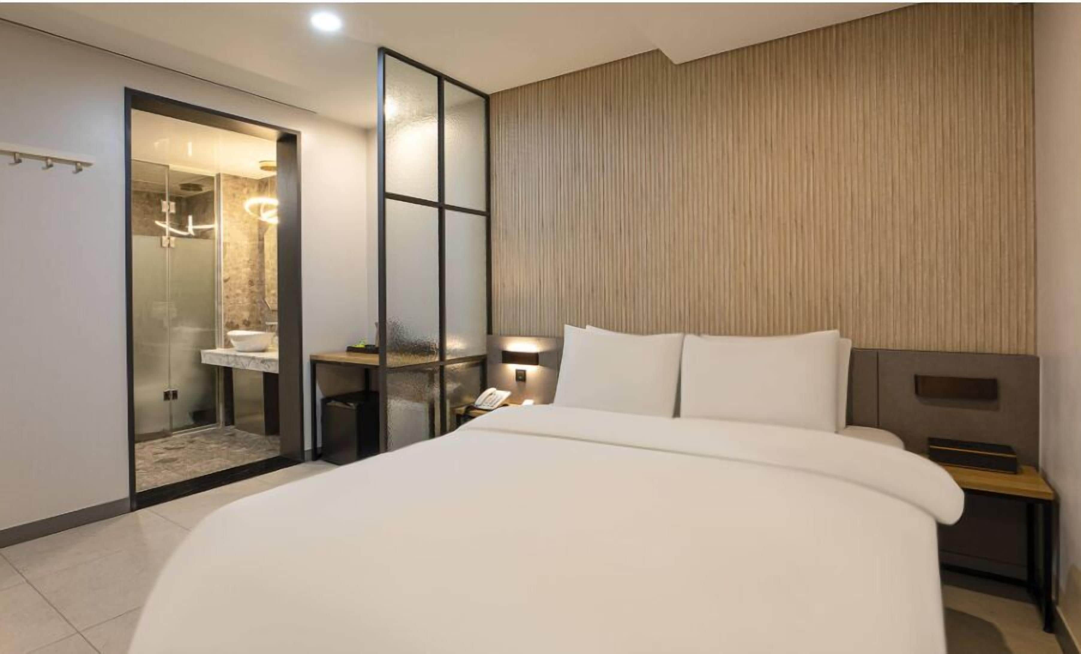 A modern hotel room with a large bed, a bedside table with a lamp, a small table with a phone, and a shower area with a glass door.