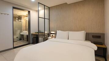A modern hotel room with a large bed, a bedside table with a lamp, a small table with a phone, and a shower area with a glass door.