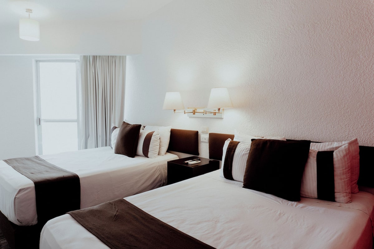 Een hotelkamer met twee bedden, een nachtkastje, een lamp en een raam met gordijnen.