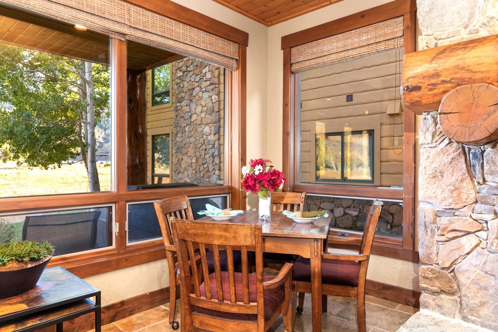 A dining area with a stone wall, wooden chairs, a table with a vase of flowers, and large windows with blinds.