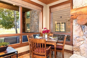 A dining area with a stone wall, wooden chairs, a table with a vase of flowers, and large windows with wicker shades. - Deer Valley Bct-501 Hot Tub KBM Resorts (Park City)