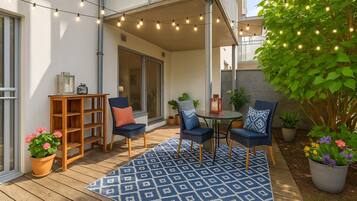An outdoor patio with a wooden deck, blue patterned rug, two blue chairs, a small round table, and string lights.