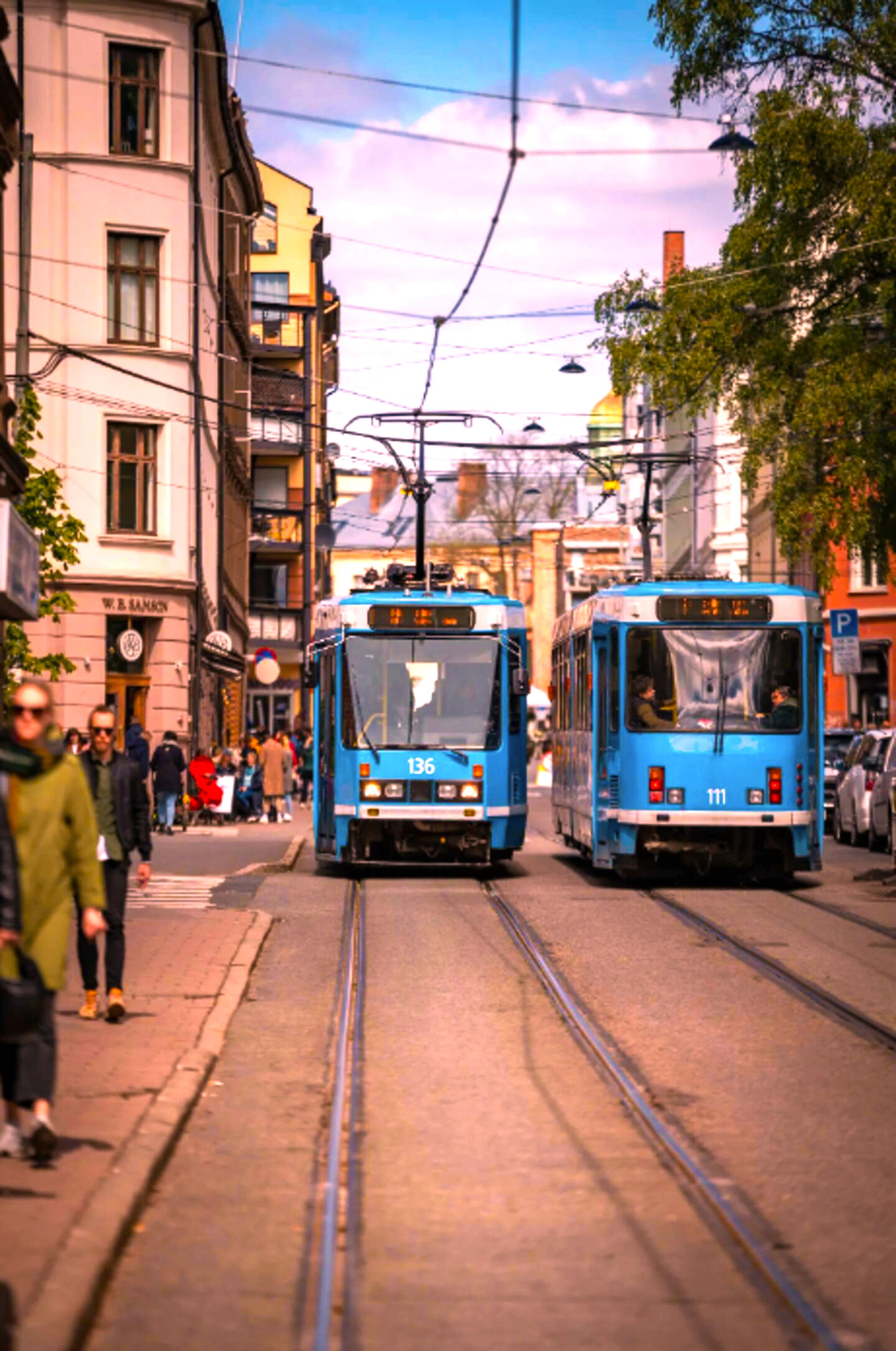 Um eléctrico nos carris numa área urbana, com edifícios e pessoas a pé.