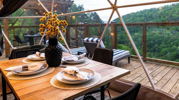A dining area with a wooden table set for two, a vase with yellow flowers, and a view of a forested hillside. - Biggest Deck! Beautiful Dome (Pikeville)