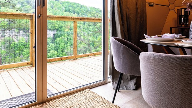 Een kamer met een groot raam dat uitkijkt op een houten terras en weelderige begroeiing. Er staat een tafel met stoelen en een koffieapparaat.