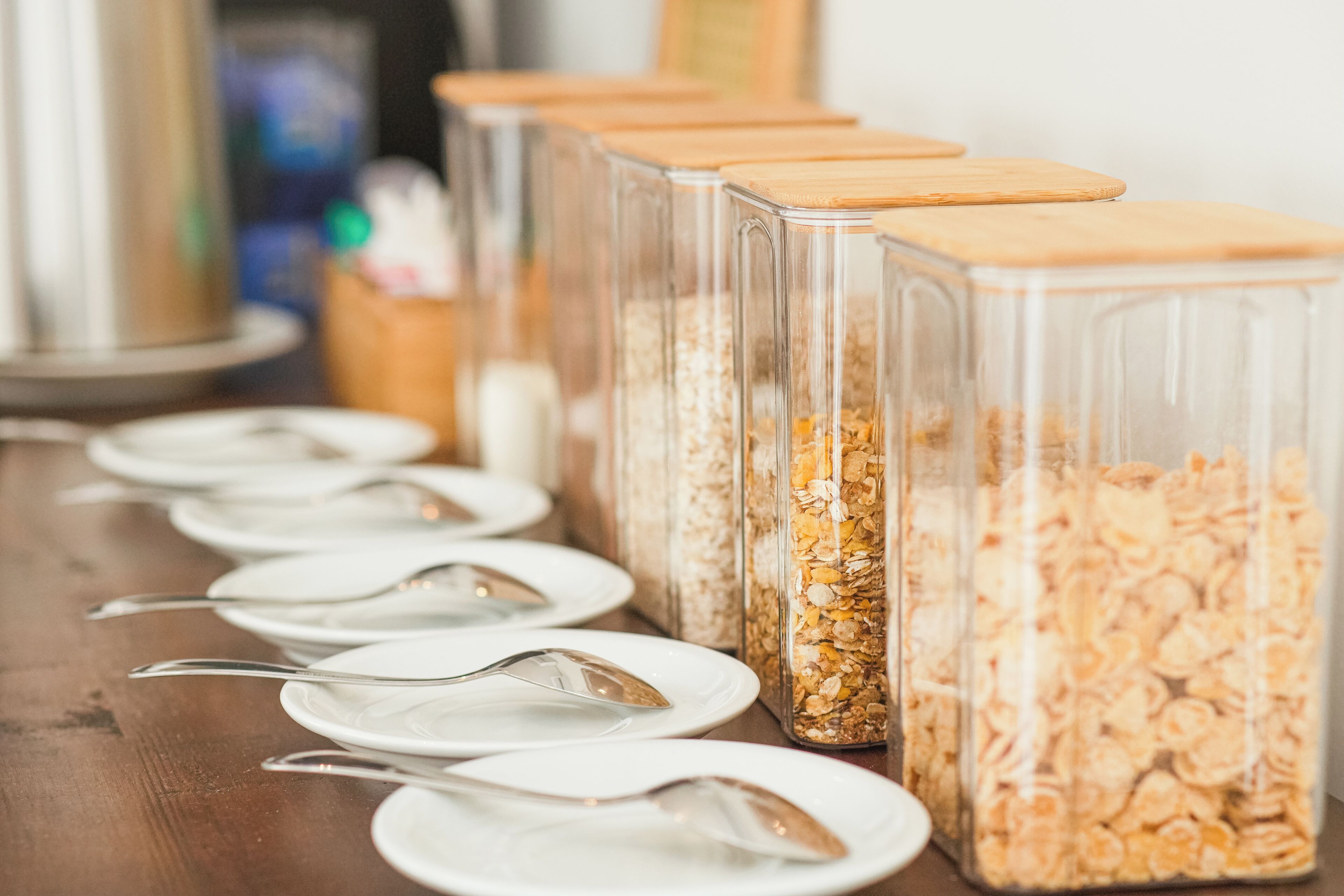A buffet setup with clear glass containers filled with cereal and topped with wooden lids, accompanied by white plates and silver spoons.