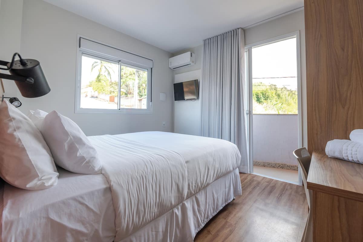 A bedroom with a bed, a TV, an air conditioning unit, a window, and a view of greenery outside.