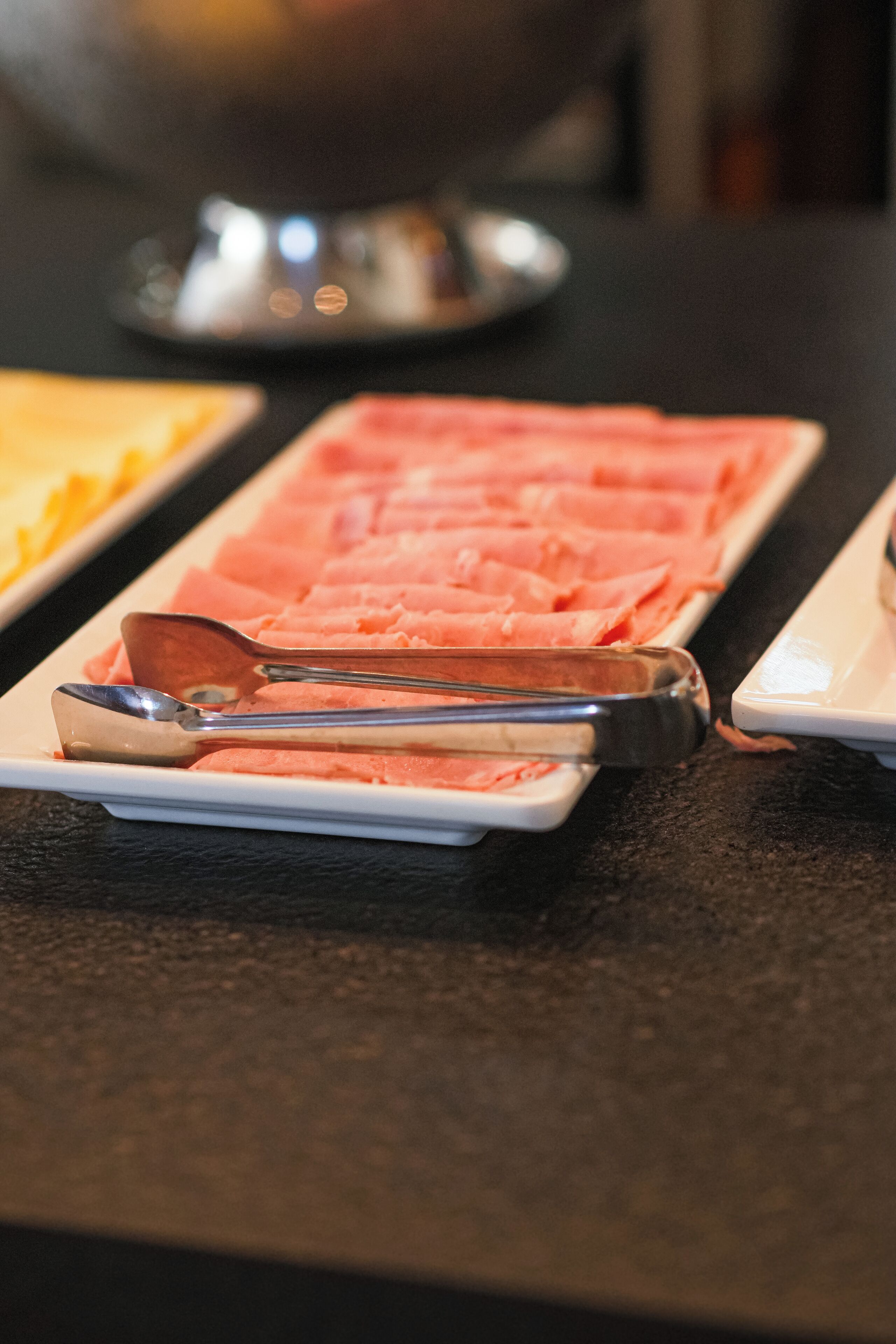 En buffetbord med pænt anrettet kød i skiver og et par tang på en hvid tallerken.
