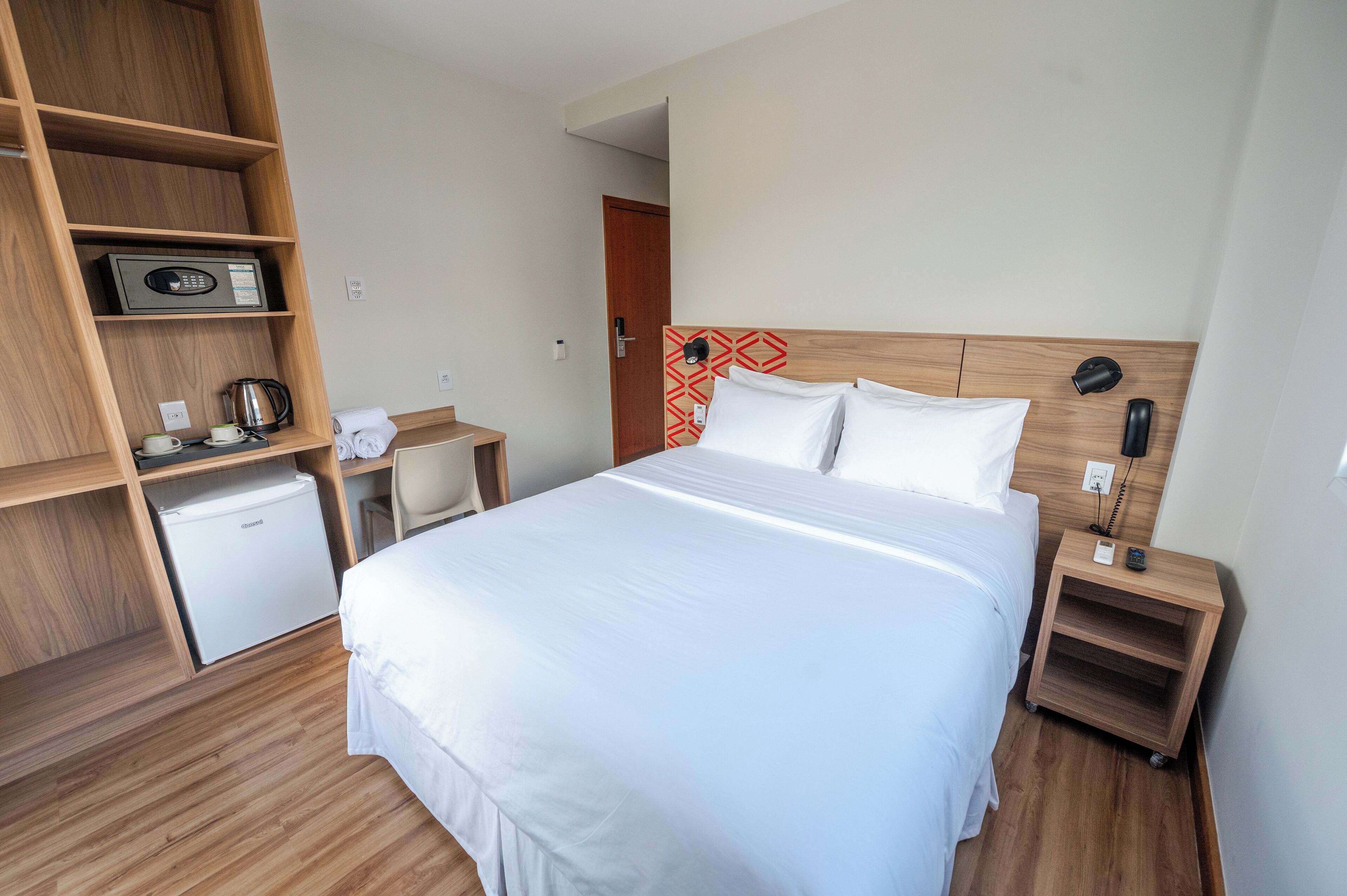 Habitación de hotel con una cama grande, un armario de madera, un escritorio pequeño con un hervidor y una mesita de noche con lámpara.