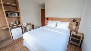 Habitación de hotel con una cama grande, un armario de madera, un escritorio pequeño con un hervidor y una mesita de noche con lámpara.