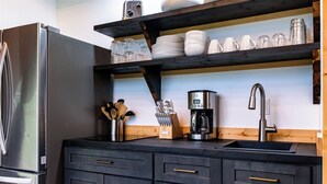 A kitchen with a stainless steel refrigerator, a coffee maker, and a sink with a faucet. There are wooden cabinets and a shelf with dishes and glasses. - Spacious Glamping Dome - Canyon Views + Hot Tub - Tennessee Wilderness (Pikeville)