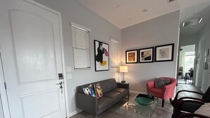 Un salon avec un canapé gris, un fauteuil rouge, une table basse en verre et une lampe. Des tableaux encadrés ornent le mur, et une porte blanche est visible.
