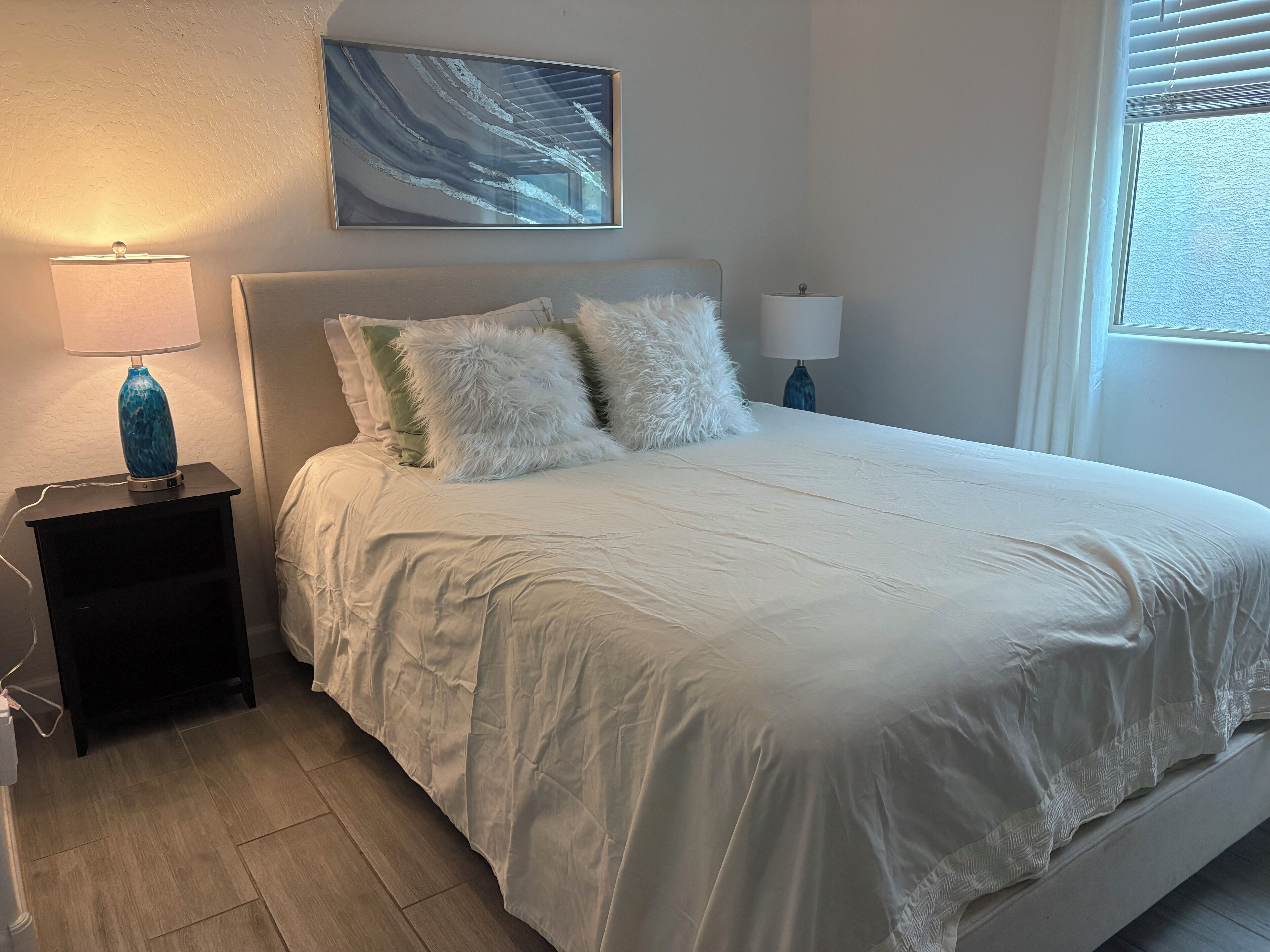 Una cama bien hecha con ropa de cama blanca, dos almohadas mullidas y un jarrón azul sobre una mesita de noche.