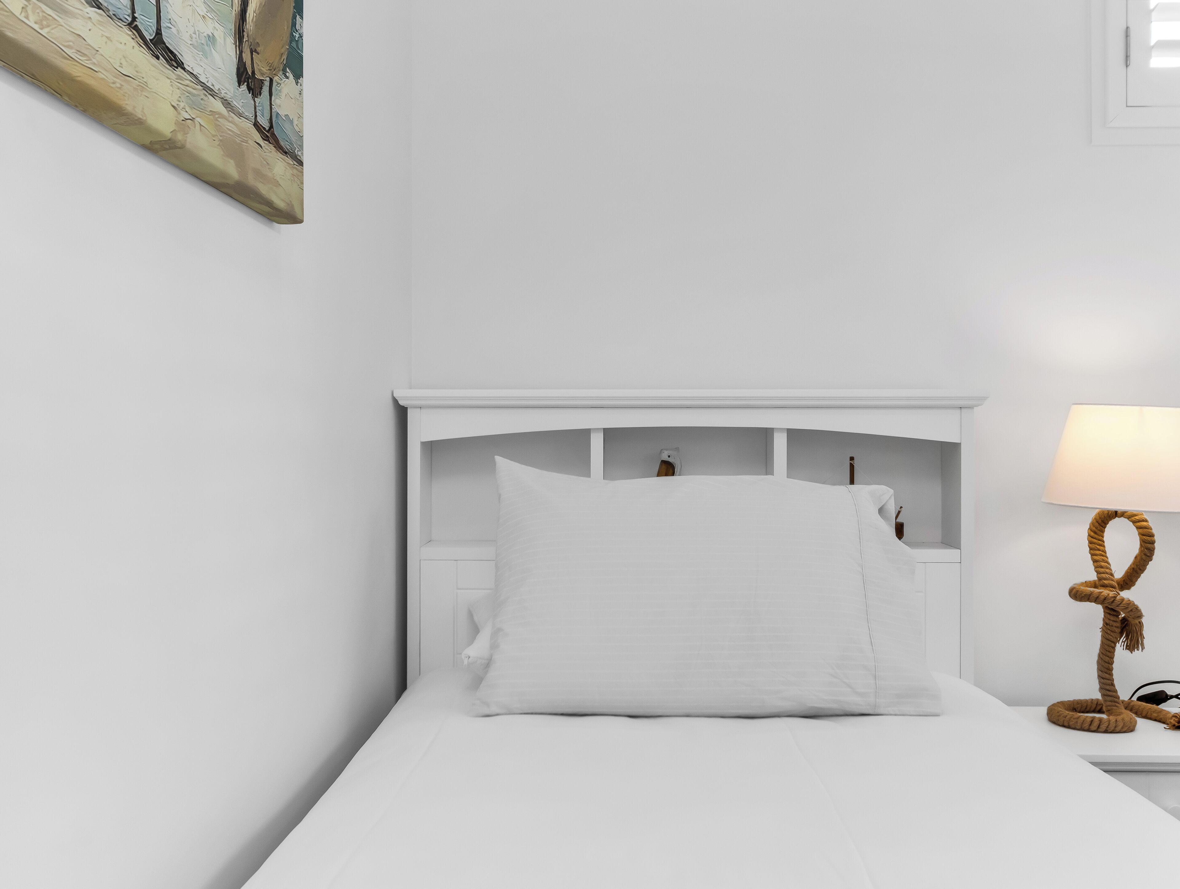 A white bed with a headboard, a bedside lamp, and a painting on the wall.