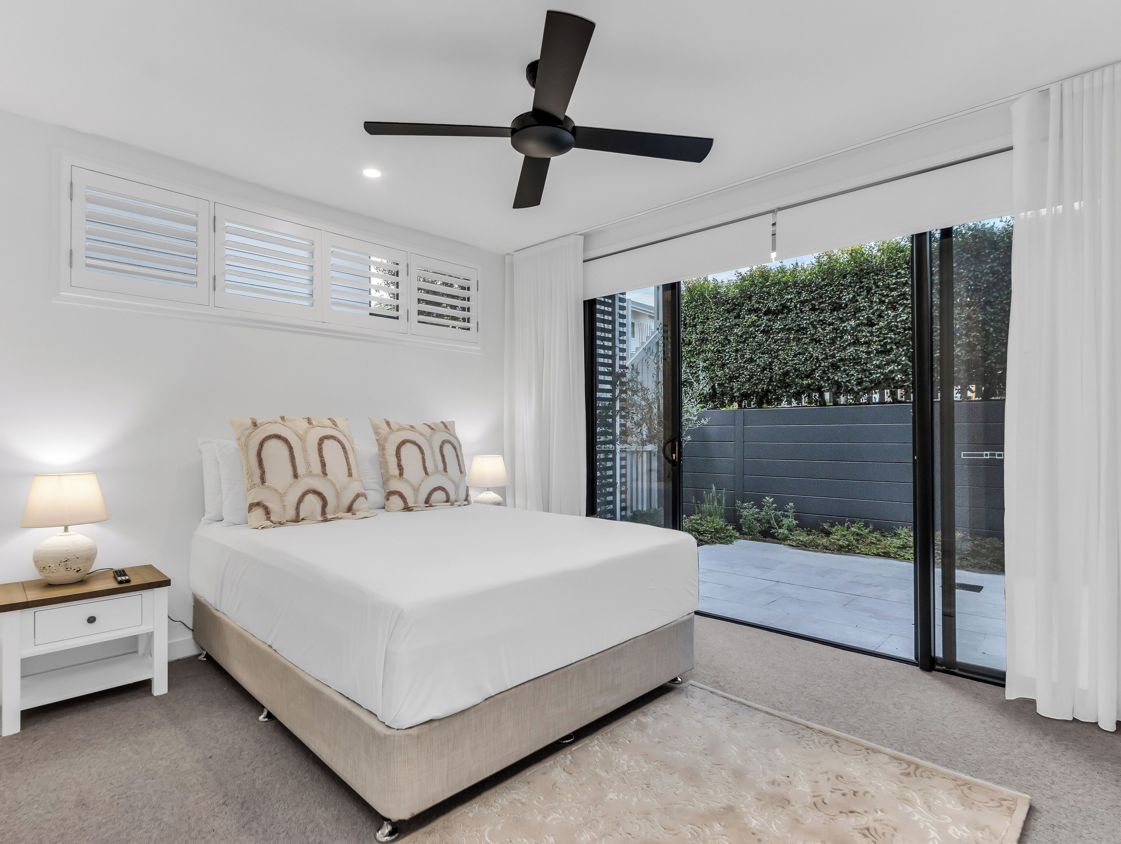 Un dormitorio con cama, lámparas de noche, ventilador de techo y vistas amplias a un espacio exterior mediante puertas correderas.