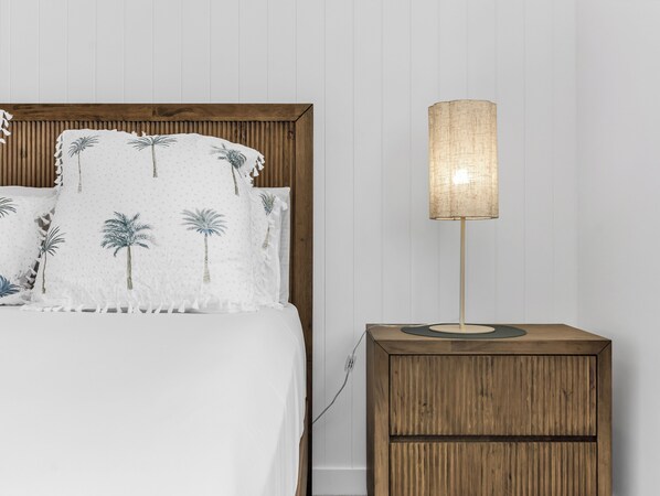 Una cama con colcha blanca adornada con motivos de palmeras, cabecera de madera y mesita de noche con lámpara.