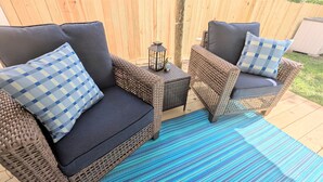 Un patio con mobiliario de mimbre, incluyendo dos sillas y una mesita, además de un tapete azul a rayas.
