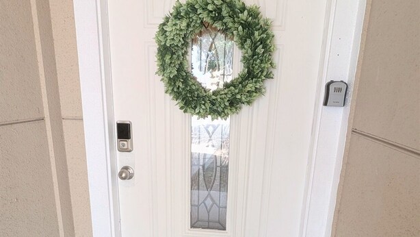 Puerta blanca con un adorno de laurel verde y un felpudo negro que dice 'Qué bueno estar en casa'.