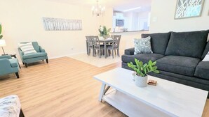 Una sala moderna con un sofá gris, sillones turquesa, una mesa de centro blanca con una planta, y una zona de comedor al fondo.