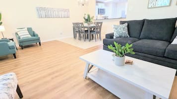 A modern living room with a grey sofa, teal armchairs, a white coffee table with a plant, and a dining area in the background.