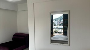 A bedroom with a bed, a small sofa, a mirror, and a window with a view of buildings and greenery. - Residencial Vista Bela Apt 09 (Two Suites) (Centro Bombinhas)