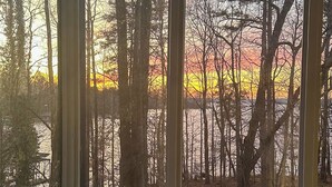 Een kamer met een groot raam dat uitkijkt over een bebost gebied tijdens de zonsondergang.