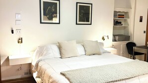 Een net opgemaakt bed met een witte deken en kussens, een nachtkastje met een lamp, en twee ingelijste foto's aan de muur.