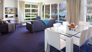 Et moderne hotelværelse med et stort vindue der giver udsigt over et smukt landskab, et hyggeligt siddeområde med sofa og lænestol, et spisebord med stole og et fladskærms-tv monteret på væggen.