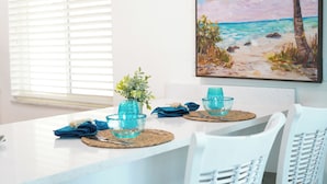 A dining area with a white table, blue glass bowls, and a painting of a beach scene.