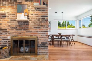 A modern living room with a brick fireplace, a dining area with a wooden table and chairs, and large windows offering a view of greenery.