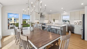 A modern kitchen with a central island, white cabinetry, and a dining area with a wooden table and chairs. - 1726 - Beach Sea Sun - Dog-Friendly Corolla Getaway | Pool & Hot Tub (Corolla)