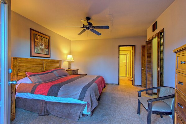 A bedroom with a bed, a chair, a ceiling fan, a nightstand, a lamp, and a door leading to another room.