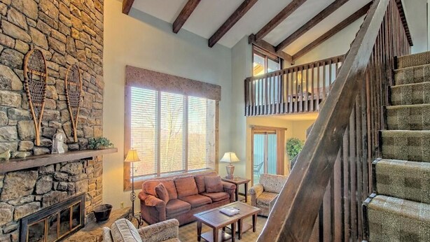 A cozy living room with a stone fireplace, a wooden staircase, and a balcony.