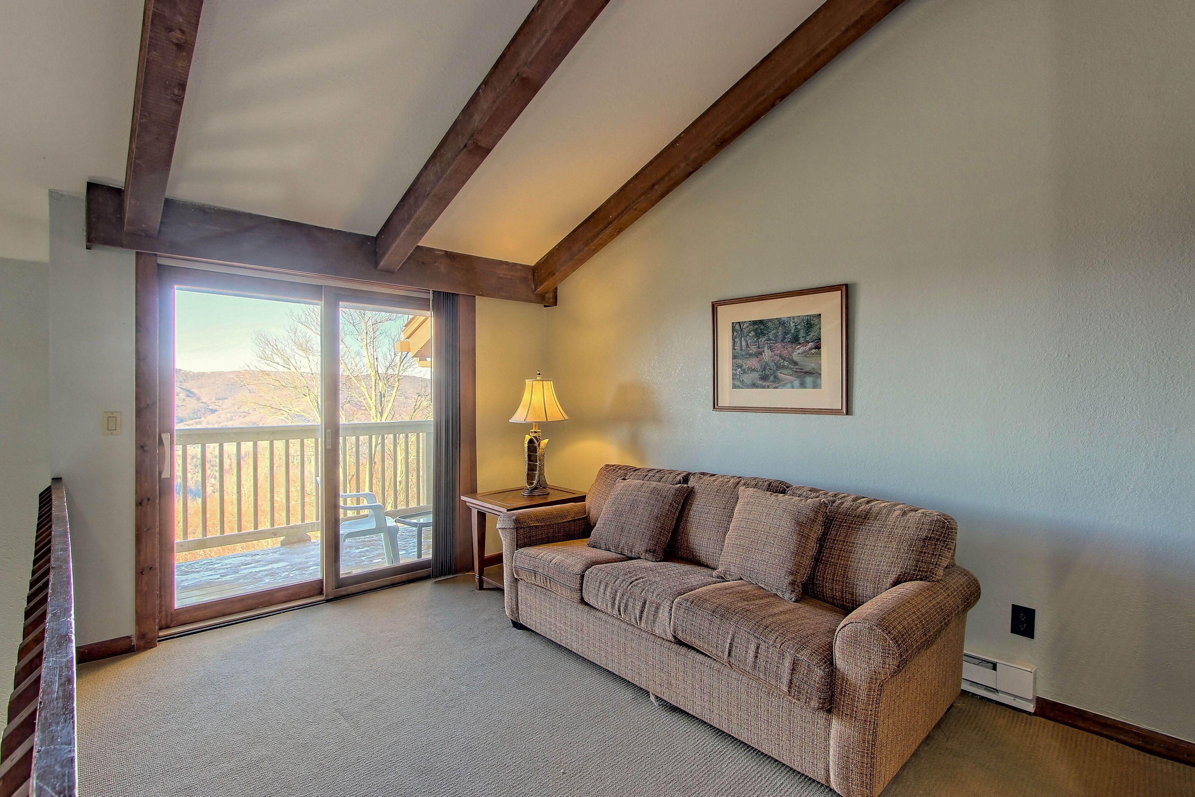 A living room with a sofa, a table with a lamp, a picture on the wall, and a sliding glass door leading to an outdoor area.