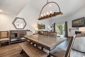 A spacious dining area with a large wooden table, comfortable seating, a decorative ceiling light, and a view of a mountain landscape through a large window.