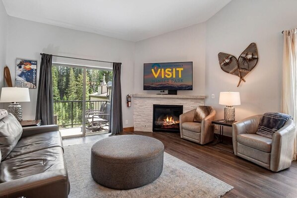 A modern living room with a fireplace, leather sofas, a round ottoman, and a large window with a view of trees.