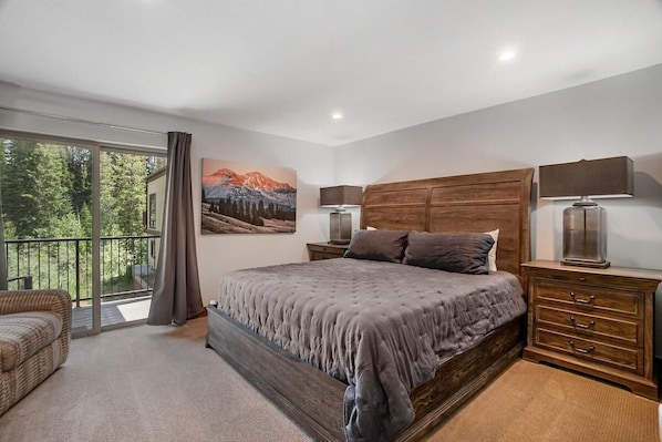 A bedroom with a large bed, a wooden headboard, a nightstand with a lamp, a dresser, and a balcony with a view of trees and mountains.