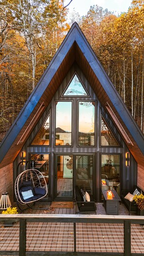 A modern cabin with a triangular roof, large windows, and a cozy seating area with a hanging chair and fire feature.