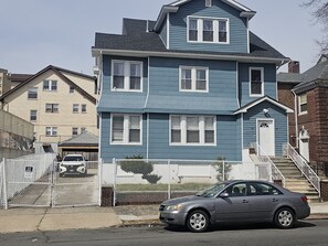 Uma casa de dois andares com fachada azul e detalhes brancos. Um carro está estacionado em frente, e uma cerca com uma placa de 'Proibido Entrar' é visível.