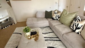 Una sala de estar con un sofá modular, una mesa de centro y un ventilador de techo. Se observan estanterías con plantas y una puerta al fondo.