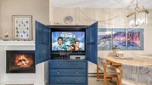 A living room with a fireplace, a television displaying 'Weekend at Bernie's,' a blue cabinet, a dining table, and a sofa. - Sunny Delighthouse – Ocean View & Beach Access (Lincoln City)