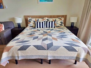 A bed with a geometric patterned bedspread, two bedside lamps, a nightstand, a gray sofa, and a framed picture on the wall.